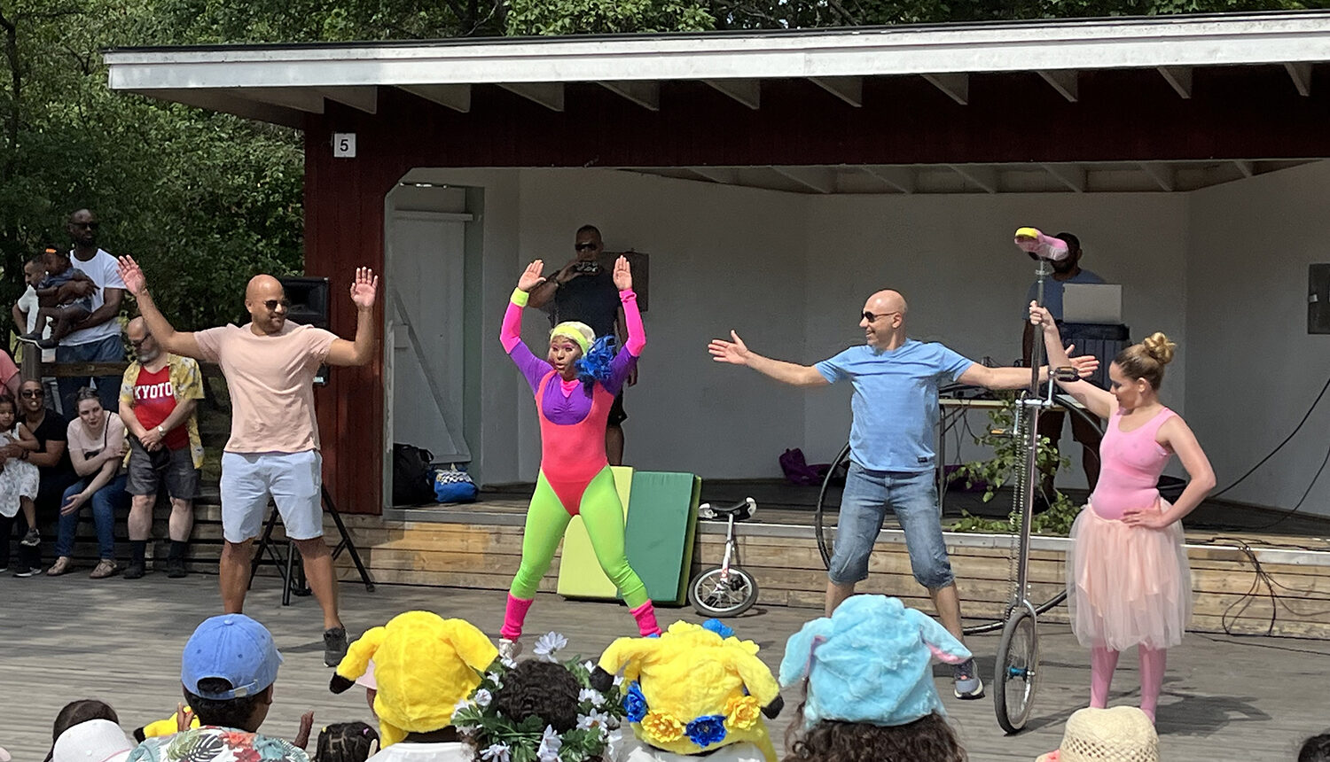 en clown i neonkläder utför dansövningar med två pappor framför barnpublik