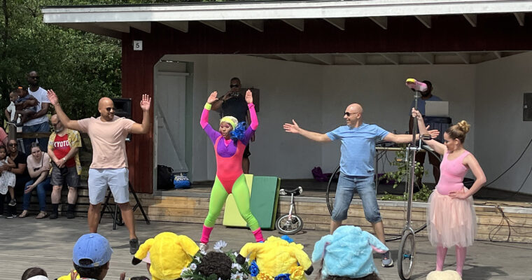 en clown i neonkläder utför dansövningar med två pappor framför barnpublik