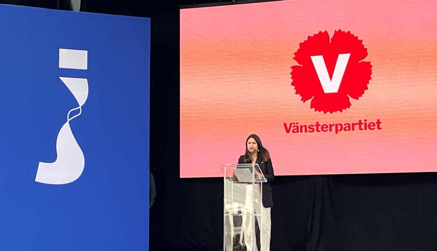 kvinna talar på stor scen framför skärmar med logotyper för Järvaveckan och Vänsterpartiet