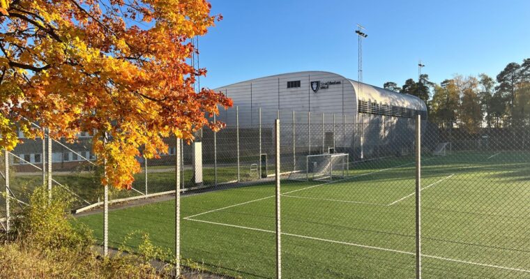 en grön fotbollsplan och en silvrig idrottshall i strålande höstväder