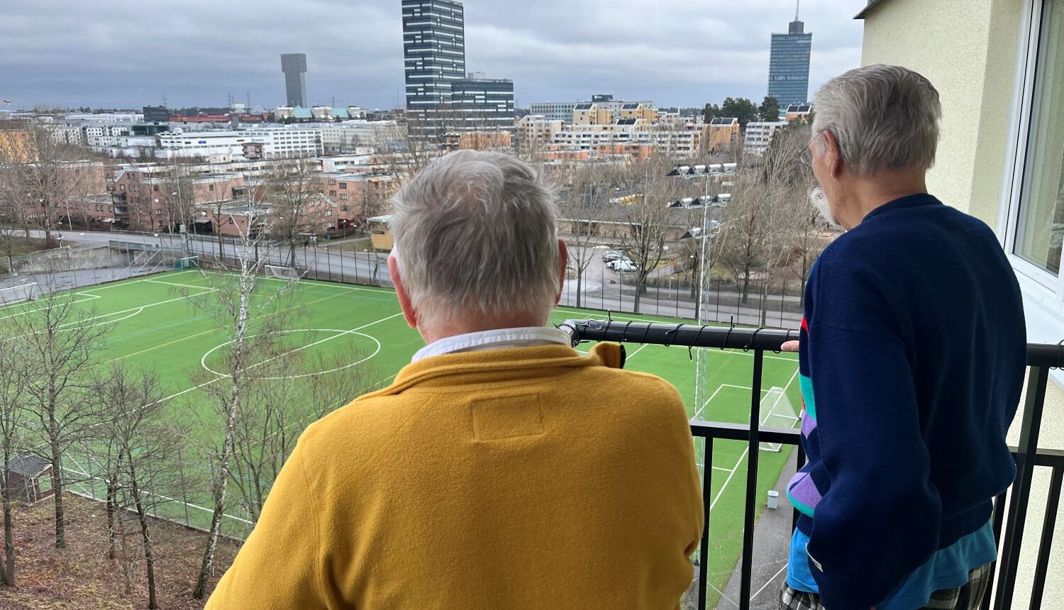 två äldre män tittar ut över en fotbollsplan och höghusvy