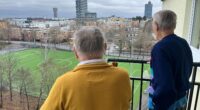 två äldre män tittar ut över en fotbollsplan och höghusvy