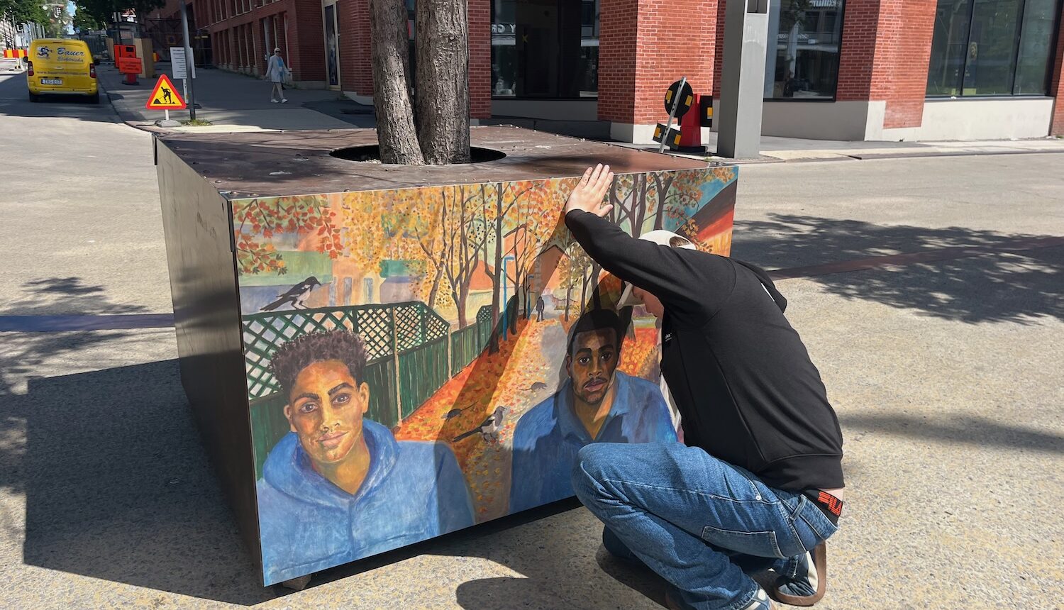 En kille sätter upp en stor affisch av en målning på en cemenkub, som ståpr runt ett träd på Stenbecks torg i Kista.