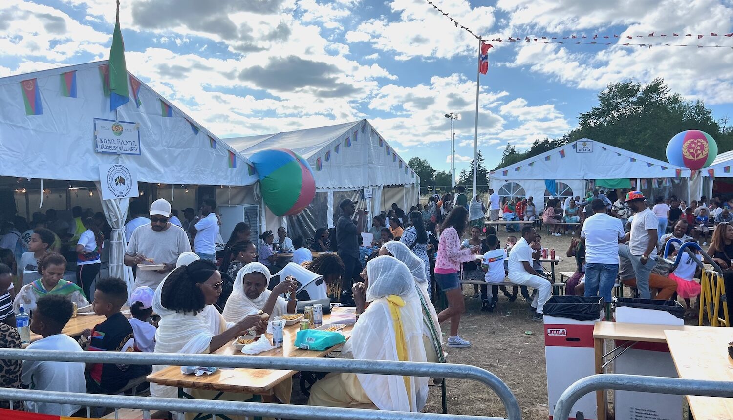 En massa människor som sitter vid bord och äter framför flera vita tält.