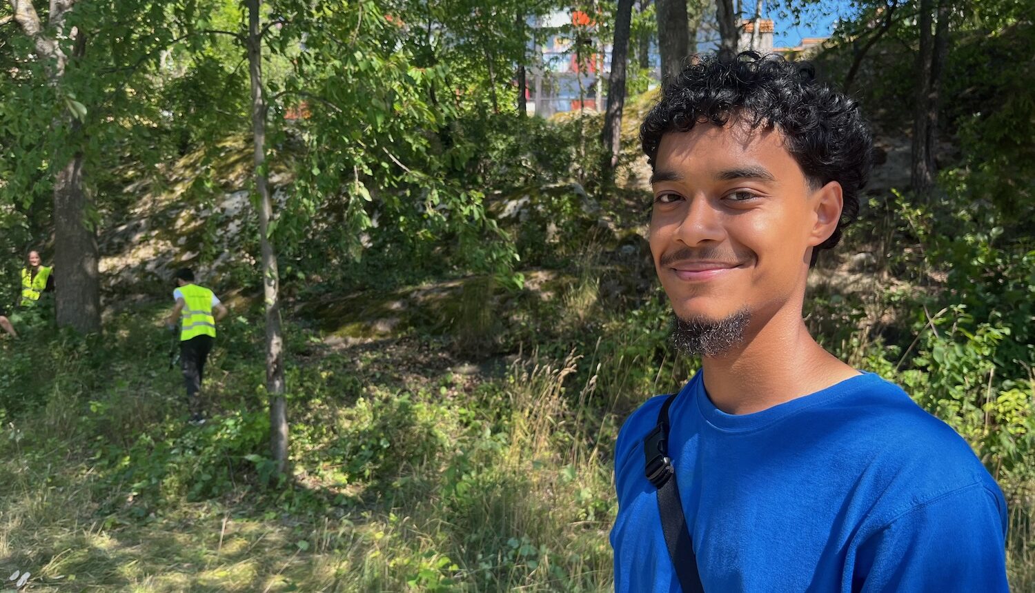 Ung man som står framför några sommarjobbare som arbetar i skogen bakom.