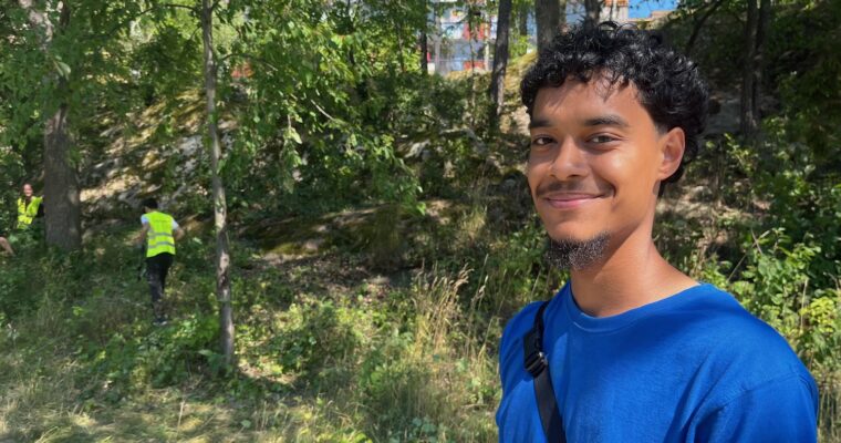 Ung man som står framför några sommarjobbare som arbetar i skogen bakom.