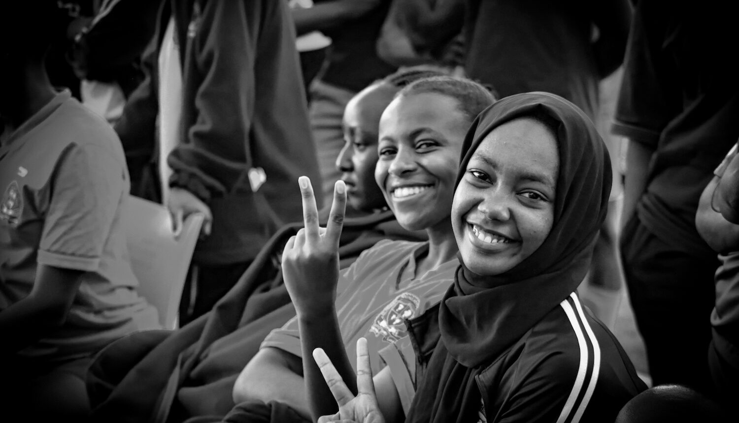 Glada tjejer, en i hijab, två utan, tittar in i kameran och gör v-tecken.