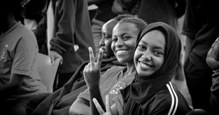 Glada tjejer, en i hijab, två utan, tittar in i kameran och gör v-tecken.