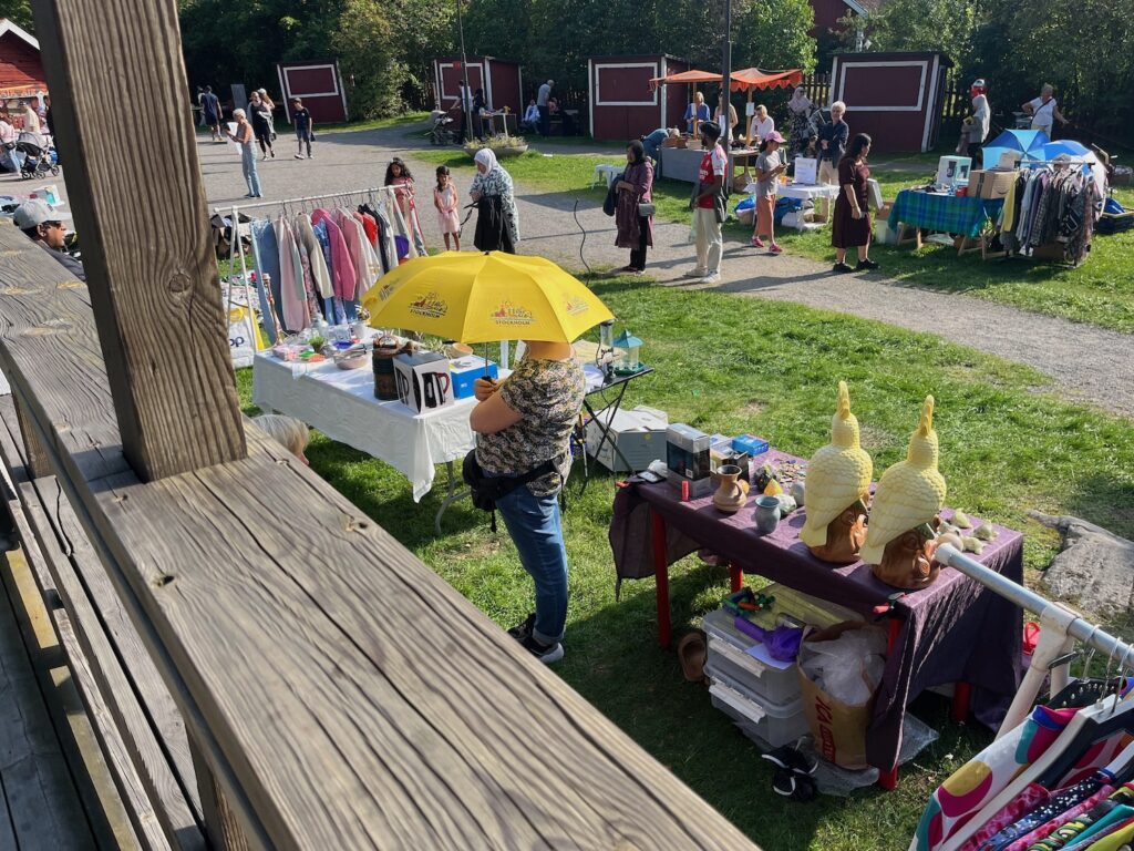 En försäljare vid ett loppisbord står under ett gult paraply, det är så varmt.