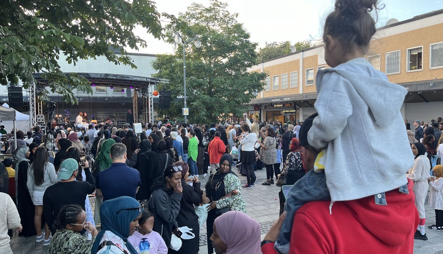 ett barn sitter på en vuxens axlar och tittar mot ett folkhav framför en scen