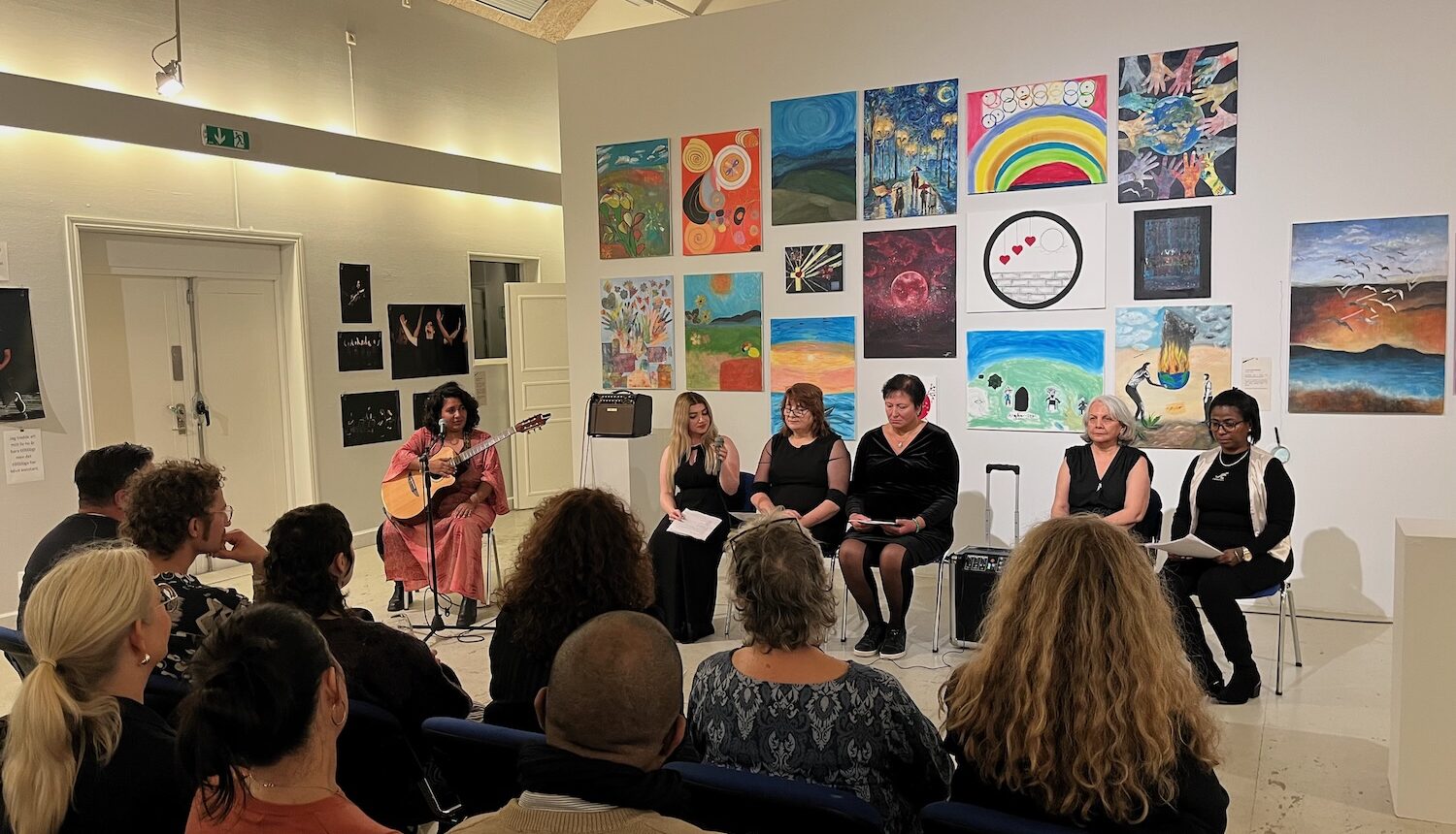 Sex kvinnor sitter framför publiken. En med en gitarr och de andra fem läser upp sina livshistorier.