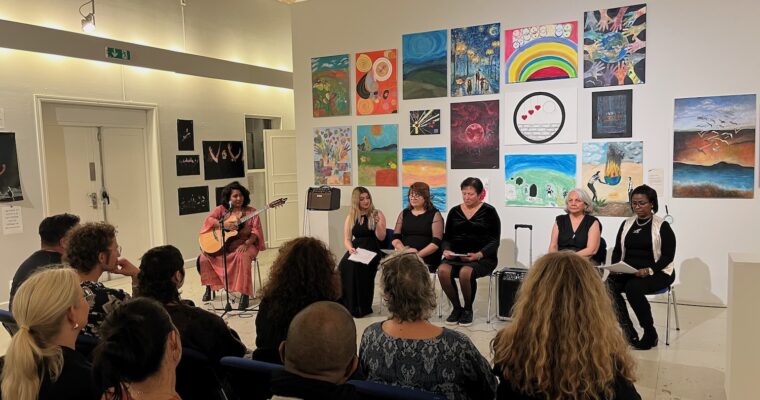 Sex kvinnor sitter framför publiken. En med en gitarr och de andra fem läser upp sina livshistorier.