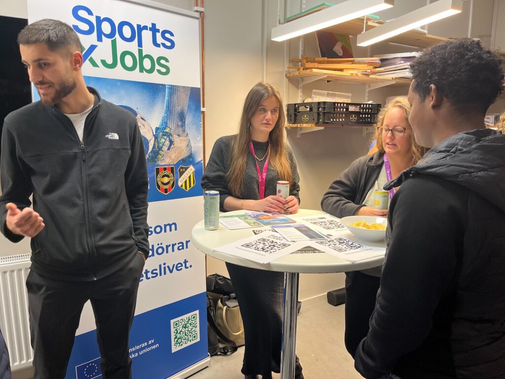 En ung man och två kvinnor vid ett hög bord. De pratar med besökare.