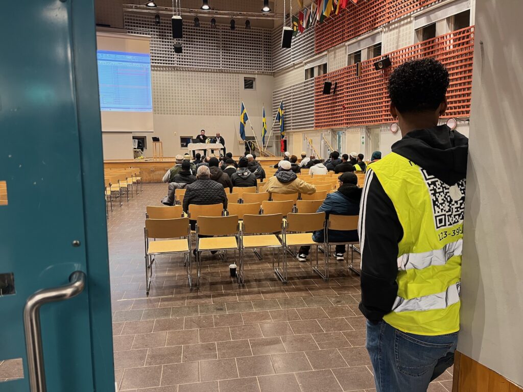 En kille i gul väst vid dörren, m¨ånga människor sitter inne i aulan.