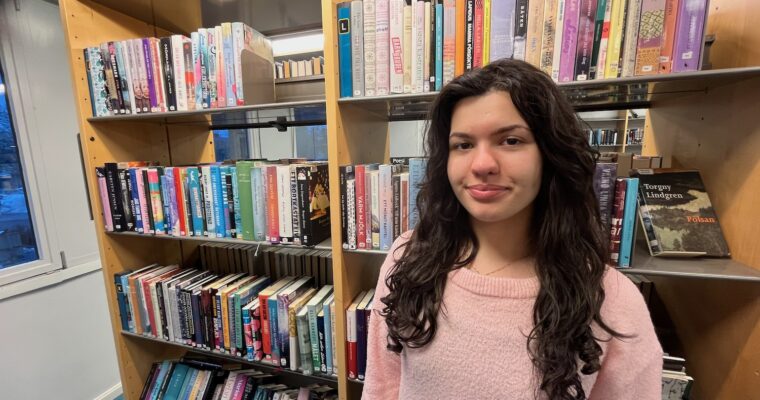 Ung tjej i långt svartlockigt hår framför bokyllor i biblioteket.