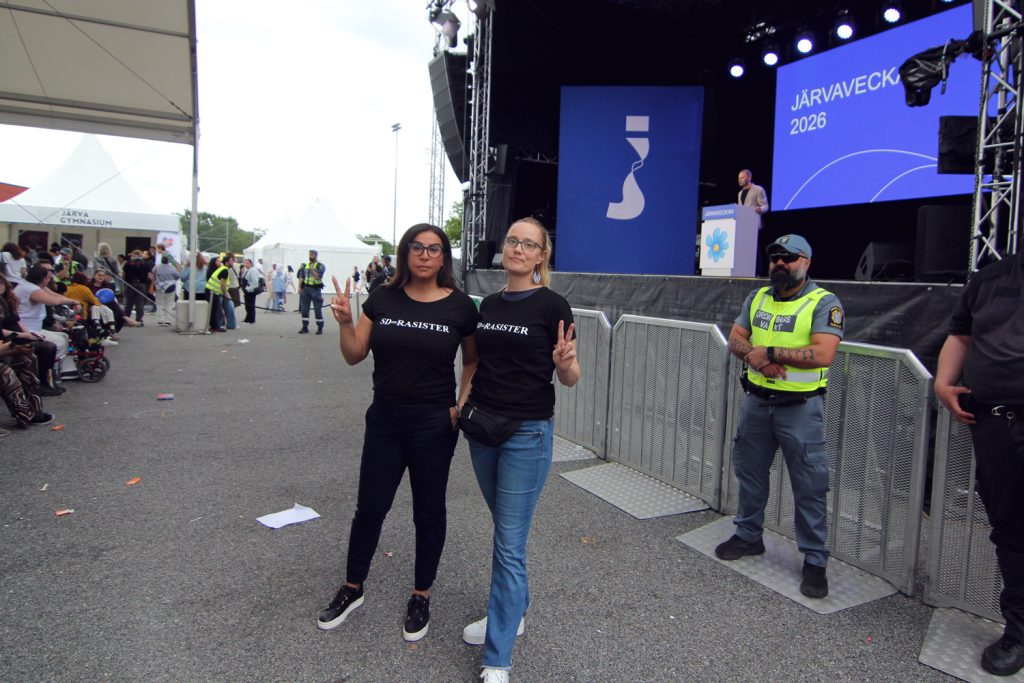 Två personer med t-shirtar med texten "SD = rasister" står framför scenen där Henrik Vinge (SD) talar.