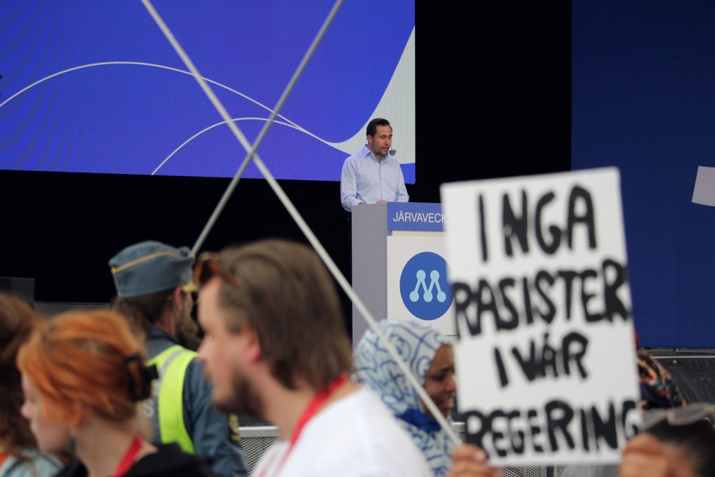 Benjamin Dousa talar på Järvaveckan. I förgrunden en skylt med texten Inga rasister i vår regering.