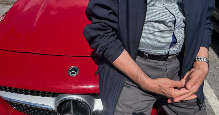 En äldre man sitter på huven till röda Mercedes Benz.