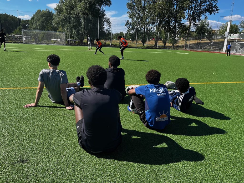 Fem grabbar som sitter på fotbollsplanen och tittar på en match. Fotograferade bakifrån.