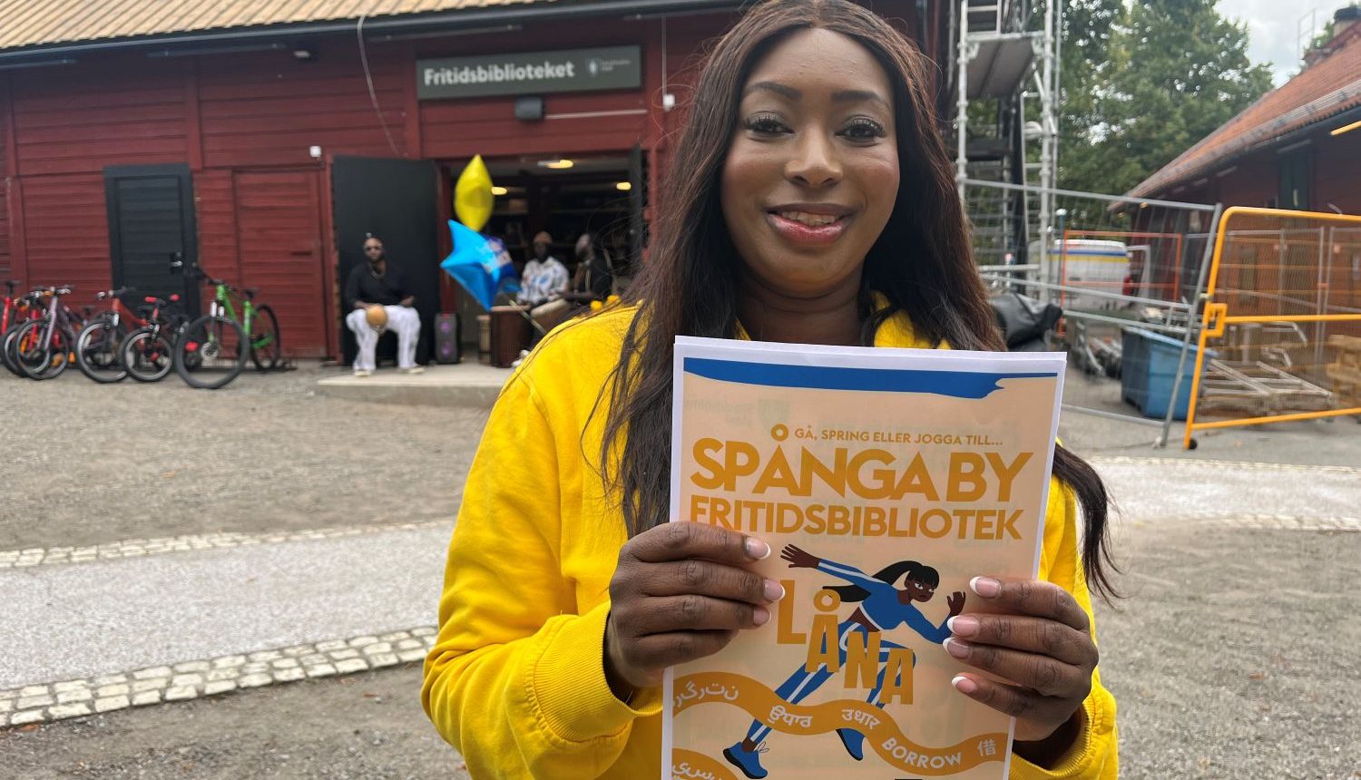 Kvinna ler mot kameran med en broschyr för Spånga by fritidsbibliotek framför sig