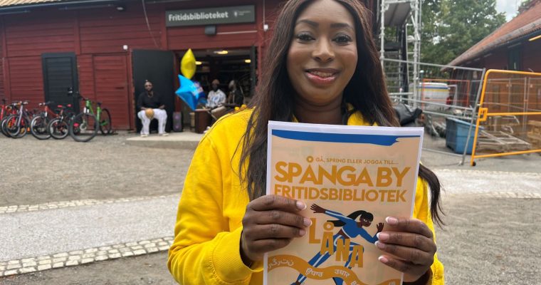 Kvinna ler mot kameran med en broschyr för Spånga by fritidsbibliotek framför sig